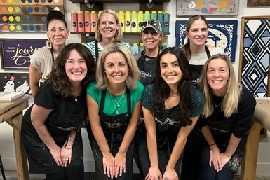 Nine smiling women in aprons pose at a lively DIY painting studio, standing in front of shelves of colorful paint bottles, wooden signs and stencils — group craft workshop photo.