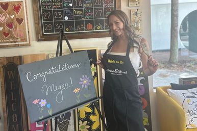 Smiling woman in a bride-to-be apron holding a paintbrush beside a chalkboard sign reading Congratulations with hand-drawn flowers in a cozy DIY craft studio during a painting party.