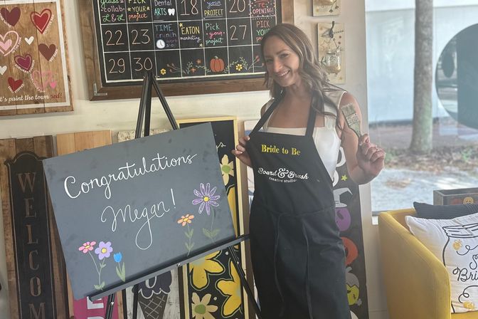 Smiling woman in a bride-to-be apron holding a paintbrush beside a chalkboard sign reading Congratulations with hand-drawn flowers in a cozy DIY craft studio during a painting party.
