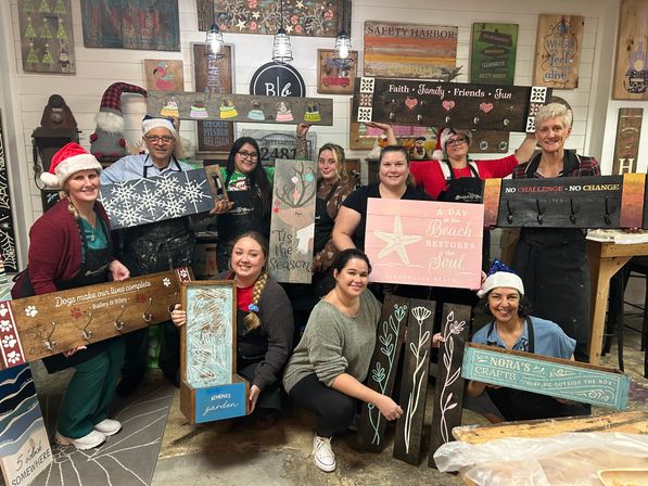 Smiling crafters in a cozy DIY wood-sign workshop wearing festive hats and aprons, proudly holding handmade painted wooden signs with holiday and beach motifs against a rustic-decor backdrop.