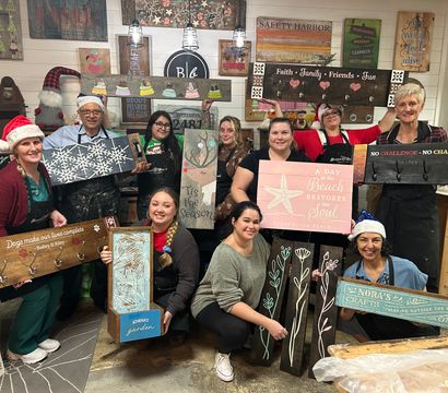 Smiling crafters in a cozy DIY wood-sign workshop wearing festive hats and aprons, proudly holding handmade painted wooden signs with holiday and beach motifs against a rustic-decor backdrop.