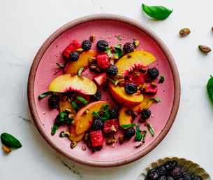 Overhead shot of a colorful summer fruit salad with sliced peaches, watermelon cubes, blackberries, chopped pistachios and fresh basil on a pink plate over white marble