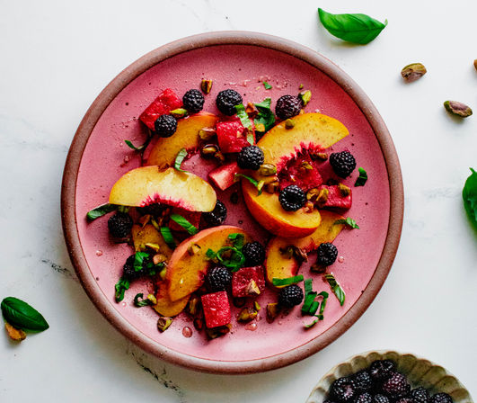 Overhead shot of a colorful summer fruit salad with sliced peaches, watermelon cubes, blackberries, chopped pistachios and fresh basil on a pink plate over white marble
