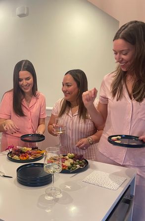 Three people in matching pink loungewear laugh around a white kitchen island, serving charcuterie boards with grapes, cheese, crackers and wine glasses for a casual indoor gathering.