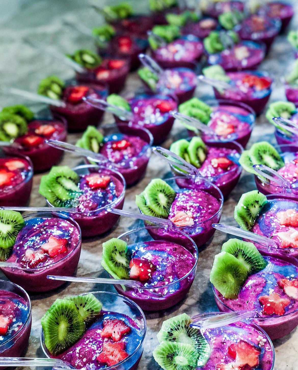 Rows of vibrant purple smoothie cups (pitaya/acai) topped with green kiwi slices and small strawberry pieces, each served in a clear plastic cup with a spoon — colorful healthy dessert display.