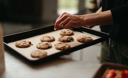 Hand sprinkling flaky salt over gooey chocolate chunk cookies on a parchment-lined baking tray in a cozy home kitchen.