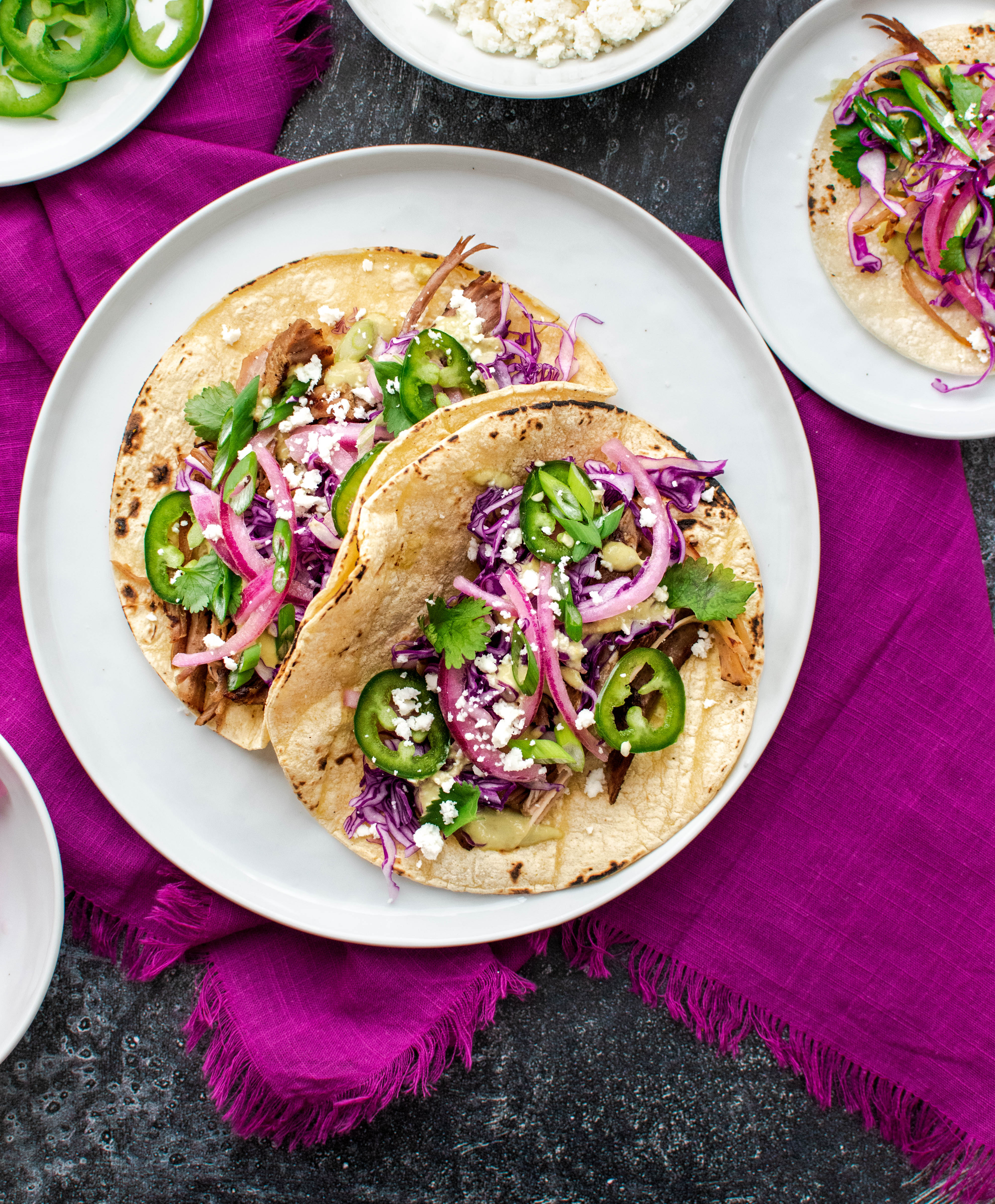 Two vibrant Mexican-style street tacos with shredded pork on charred corn tortillas, topped with purple cabbage, pickled red onion, sliced jalapeño, cilantro and crumbled queso, plated on a white dish over a magenta cloth.