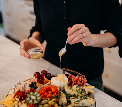 Person drizzling honey over a loaded charcuterie board on a marble kitchen counter — cheese platter with cured meats, olives, artichokes, cherries, crackers and fresh herbs.