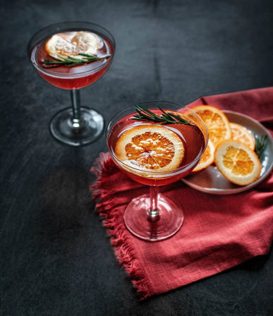 Two ruby-red citrus cocktails in coupe glasses garnished with dried orange slices and rosemary, set on a dark tabletop with a red linen napkin and a small plate of extra orange slices.