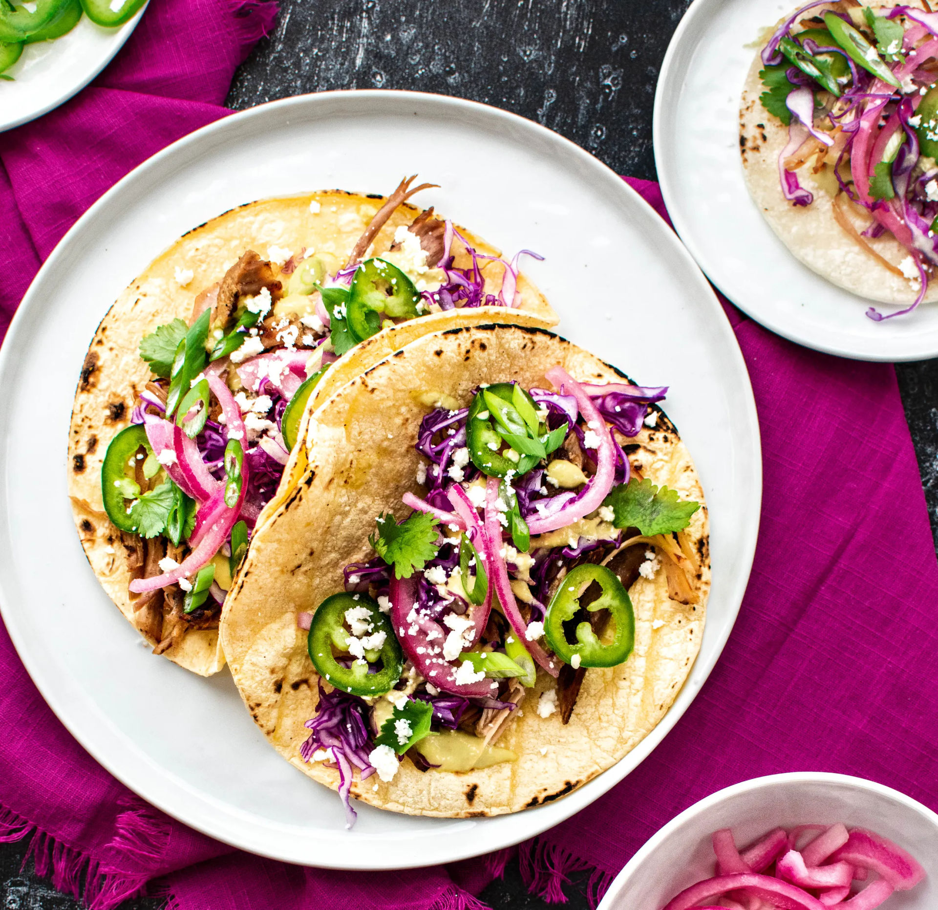 Colorful Mexican-style corn tacos with shredded meat, purple cabbage slaw, pickled red onions, jalapeño slices, cilantro and queso fresco on a white plate