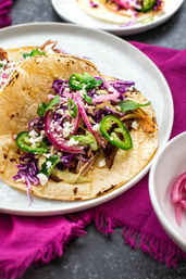Street-style Mexican taco on a charred corn tortilla topped with shredded meat, pickled red onions, purple cabbage, sliced jalapeño, cilantro and crumbled cotija cheese
