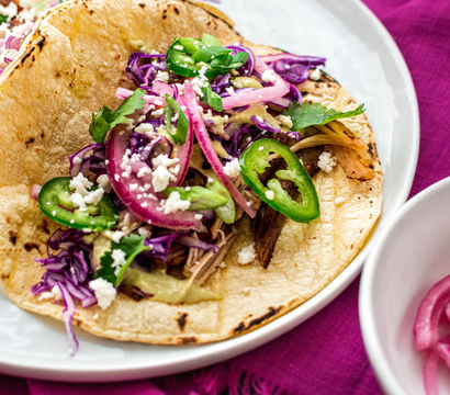 Street-style Mexican taco on a charred corn tortilla topped with shredded meat, pickled red onions, purple cabbage, sliced jalapeño, cilantro and crumbled cotija cheese