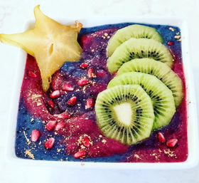 Vibrant acai-style smoothie bowl in a white square bowl topped with sliced kiwi, starfruit, pomegranate arils and edible gold dust — colorful tropical breakfast