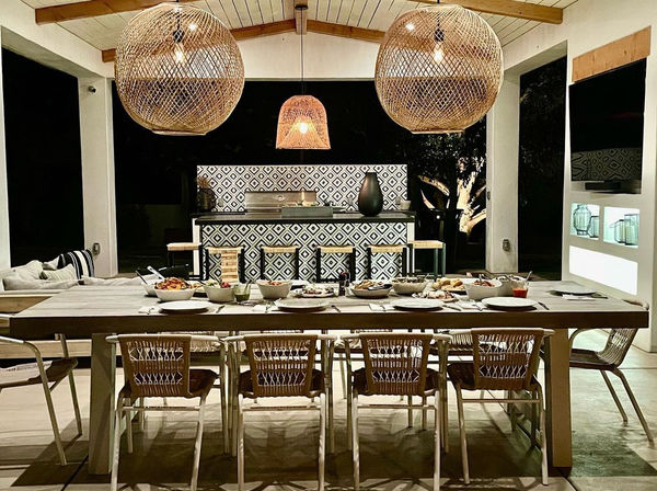 Nighttime backyard covered patio and outdoor dining area with a long wooden table set for dinner, woven rattan pendant lights, wicker chairs, and a patterned-tile bar with stools and grill.