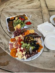 Colorful charcuterie grazing board on a rustic outdoor wooden table — mixed cheeses, cured meats, grapes, olives, dried apricots, crackers and two dips, with stacked plates and napkins for a casual patio gathering.