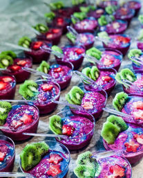 Rows of vibrant purple smoothie cups (pitaya/acai) topped with green kiwi slices and small strawberry pieces, each served in a clear plastic cup with a spoon — colorful healthy dessert display.