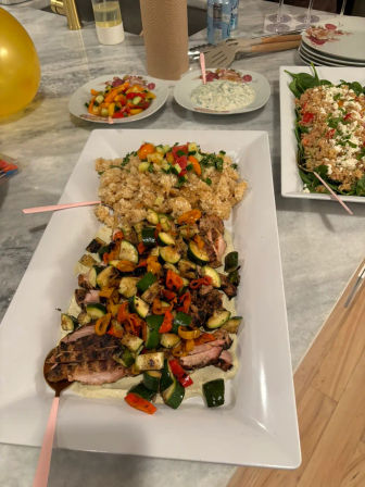 Platter of sliced grilled meat topped with roasted zucchini and colorful bell peppers next to a mound of seasoned rice and several side salads on a marble kitchen island — colorful home buffet.