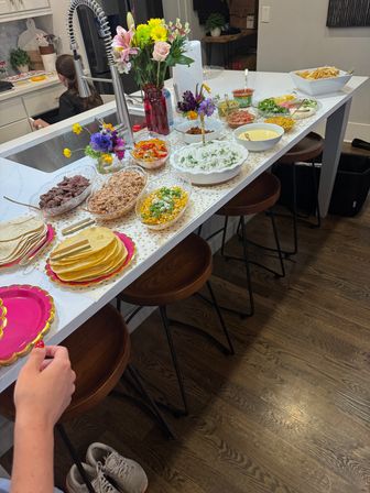 Home kitchen island taco bar on a white countertop with stacks of tortillas, grilled meat, shredded chicken, cilantro rice, corn salad, salsas, chips and a colorful vase of flowers
