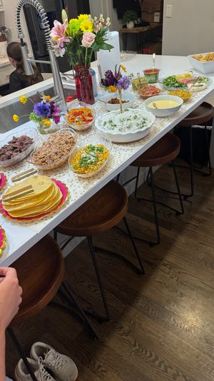 Home kitchen island taco bar on a white countertop with stacks of tortillas, grilled meat, shredded chicken, cilantro rice, corn salad, salsas, chips and a colorful vase of flowers