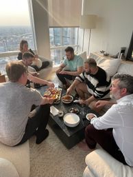 Six friends enjoying a charcuterie board, bread and wine around a low coffee table in a sunlit high-rise apartment living room with floor-to-ceiling city skyline views.