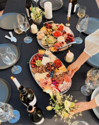 Elegant outdoor dining table on a black tablecloth with two colorful charcuterie boards — cheeses, cured meats, crackers, olives, cherries, blueberries, kiwi, nuts and dips — surrounded by wine bottles, stemmed glasses, lit candles and small floral arrangements; hands arranging a board.