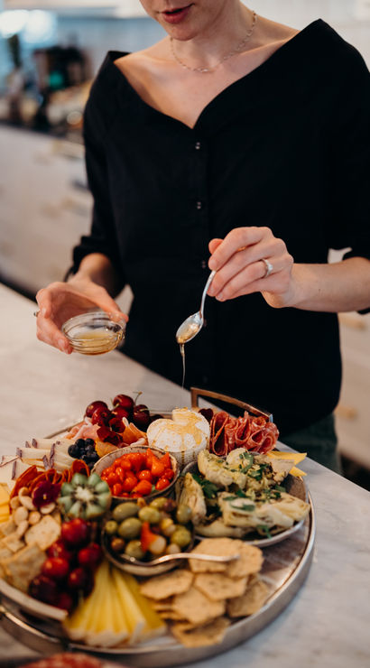 Hands drizzling honey onto a soft cheese on a vibrant charcuterie board with cured meats, olives, marinated artichokes, cherries, cherry tomatoes, assorted cheeses and crackers on a marble kitchen countertop.