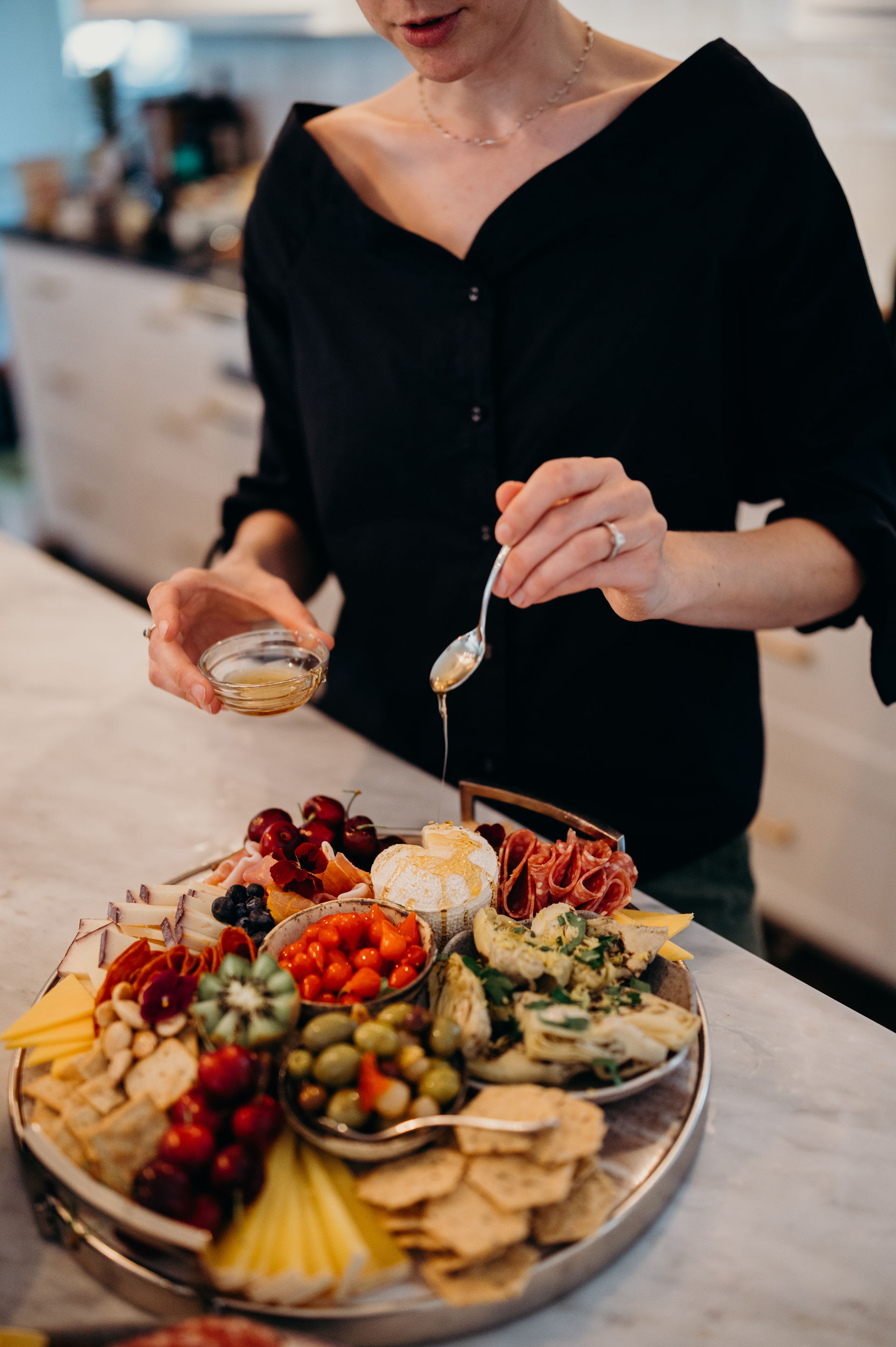 Hands drizzling honey onto a soft cheese on a vibrant charcuterie board with cured meats, olives, marinated artichokes, cherries, cherry tomatoes, assorted cheeses and crackers on a marble kitchen countertop.