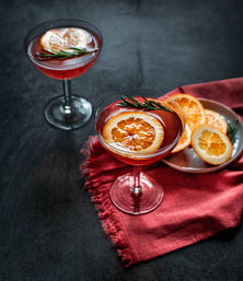 Two red cocktails in coupe glasses garnished with dried orange slices and rosemary, plate of extra orange slices on a red cloth over a dark tabletop
