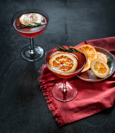 Two red cocktails in coupe glasses garnished with dried orange slices and rosemary, plate of extra orange slices on a red cloth over a dark tabletop