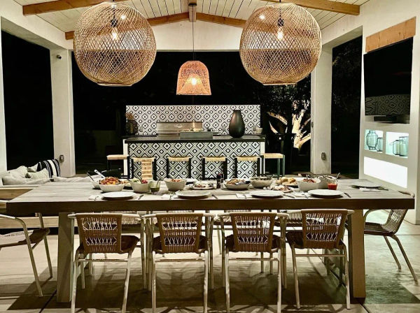 Covered outdoor dining patio at night with a long wooden table set for dinner, woven rattan pendant lights, patterned tile bar island with stools, and relaxed modern‑boho seating and ambient lighting.