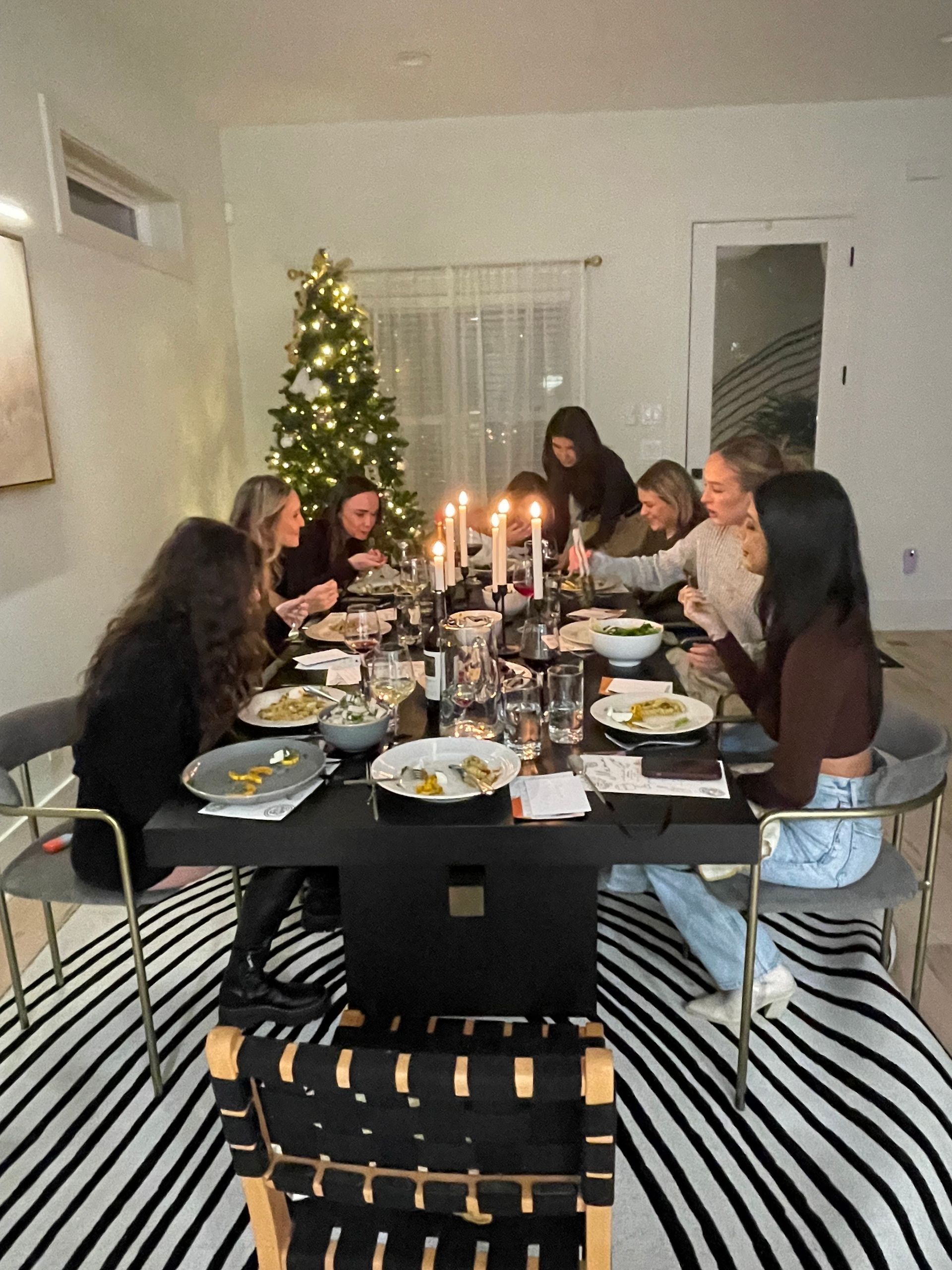 Cozy candlelit holiday dinner with a group of friends seated around a modern black dining table, plates and wine glasses on the table and a lit Christmas tree glowing in the contemporary living room.