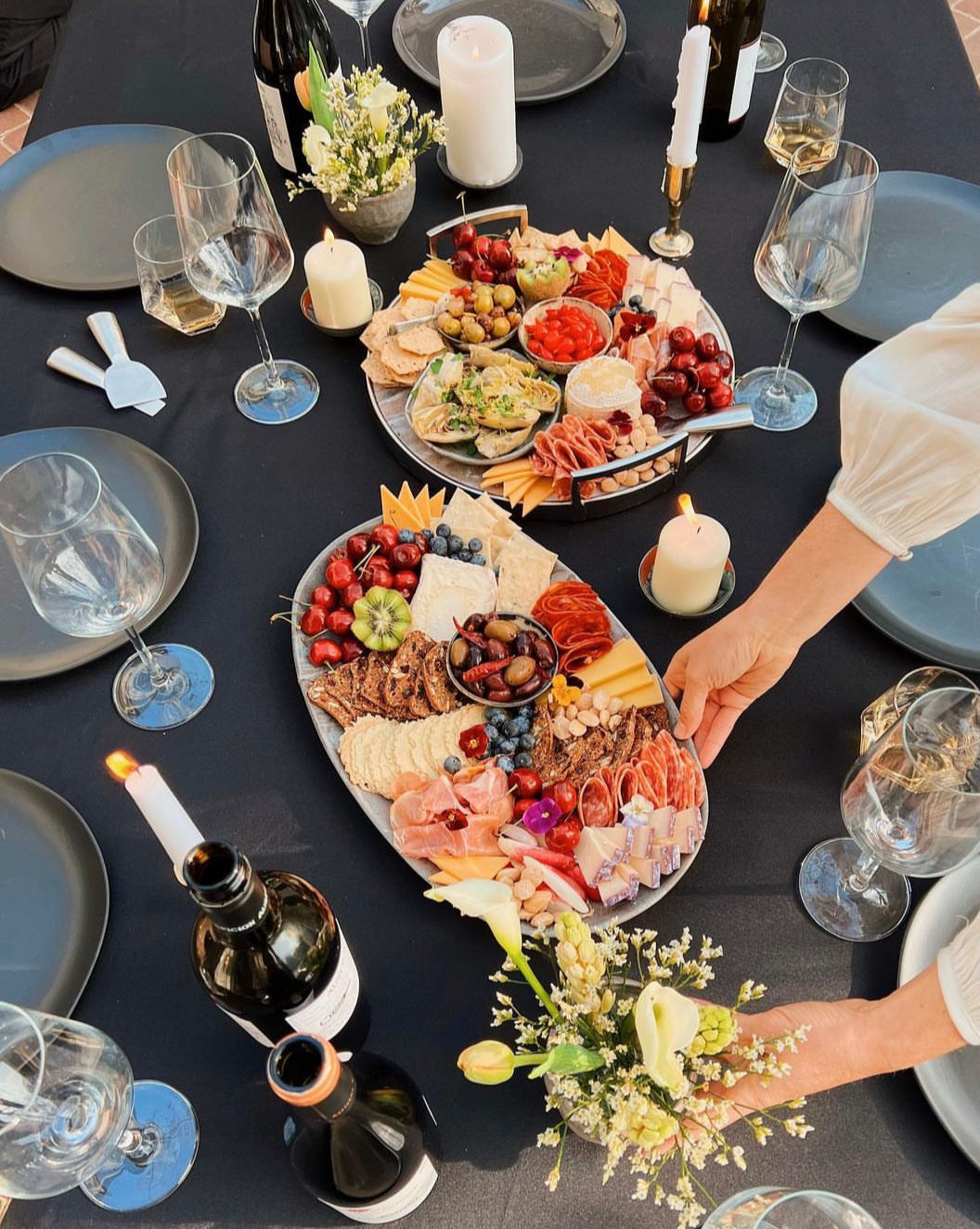 Inviting outdoor tablescape with two charcuterie platters of cheeses, cured meats, olives, crackers, cherries and berries, surrounded by wine bottles, glasses, lit candles and small floral arrangements on a black tablecloth.