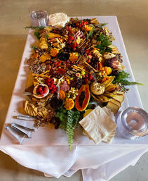 Overhead view of a colorful indoor grazing table for an event: large charcuterie spread with assorted cheeses, cured meats, crackers, breads, olives, dips, nuts and fresh fruit (papaya, berries, figs, grapes) arranged on a white-clothed catering table.