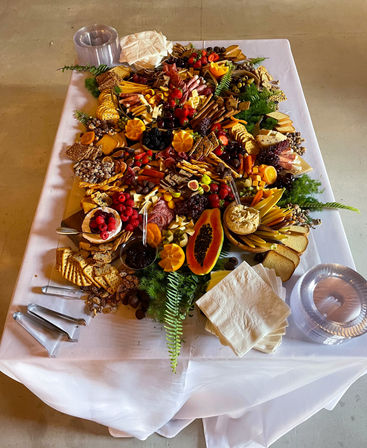 Overhead view of a colorful indoor grazing table for an event: large charcuterie spread with assorted cheeses, cured meats, crackers, breads, olives, dips, nuts and fresh fruit (papaya, berries, figs, grapes) arranged on a white-clothed catering table.
