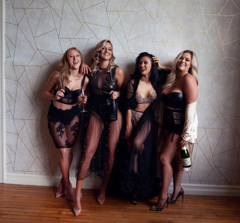 Four women laughing in lingerie and sheer boudoir outfits, holding champagne bottles and a glass in a celebratory indoor photoshoot against geometric wallpaper and hardwood floor.