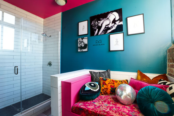 Bright modern loft bathroom with glass-enclosed white-tile shower, teal accent wall with framed black-and-white art, and a vibrant pink daybed covered in floral fabric, colorful throw pillows and a disco ball.