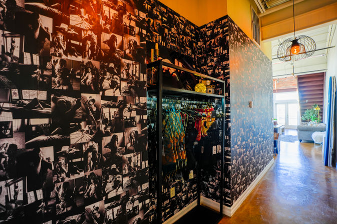 Industrial loft hallway with black-and-white photo-collage wallpaper, open clothing rack of colorful swimsuits and activewear, wire-cage pendant light and polished concrete floor leading to a sunlit living area with plants.