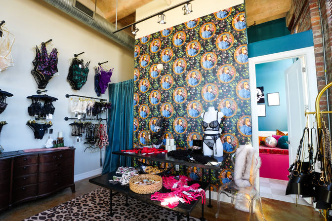 Lingerie boutique interior in a loft-style retail space with colorful patterned wallpaper, mannequin wearing a black lace set, wall racks of bras and panties, display tables with pink and red intimates, teal fitting-room curtain, leopard-print rug and exposed ductwork.