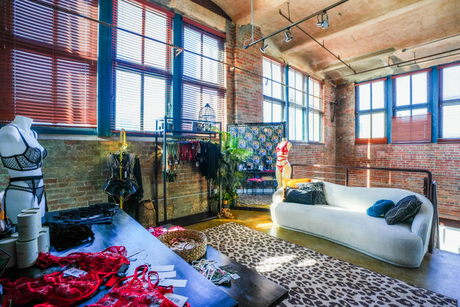 Sunlit industrial loft lingerie boutique with exposed brick walls, tall paned windows and arched concrete ceiling, mannequins and racks of colorful lingerie, plush white sofa and leopard-print rug.