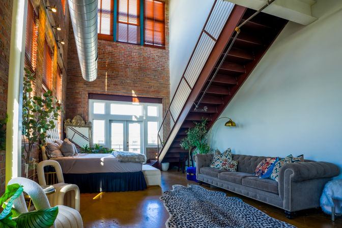 Sunny industrial loft with exposed brick walls, tall windows and metal ductwork, a steel staircase to a mezzanine, cozy bed, tufted gray sofa with colorful pillows, indoor plants and patterned rug.