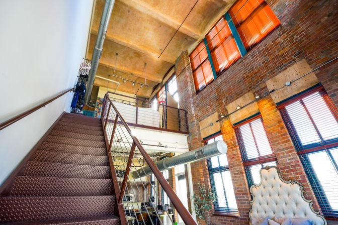 Bright industrial urban loft with metal diamond-plate staircase to a mezzanine, exposed brick walls, high ceilings, large windows with orange blinds, visible ductwork and tufted headboard