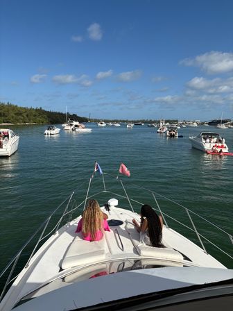 Two people relaxing on the bow of a white yacht anchored among many motorboats in a sunny coastal bay with blue sky, scattered clouds and a tree-lined shore