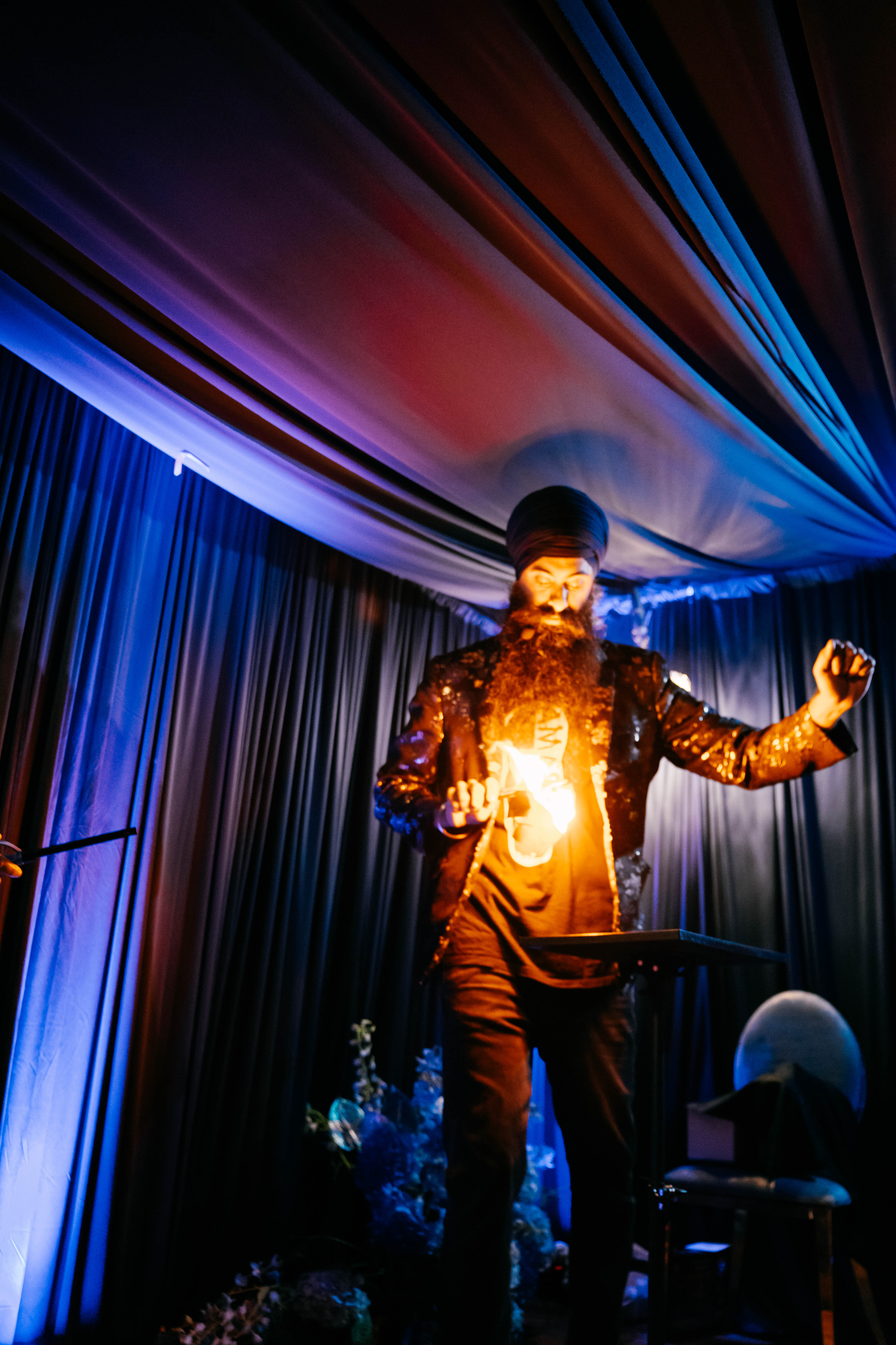 Stage performer with turban and long beard doing a fire trick while wearing a sequined jacket under dramatic blue and purple draped ceiling and curtains at an indoor event