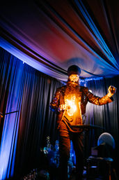 Stage performer with turban and long beard doing a fire trick while wearing a sequined jacket under dramatic blue and purple draped ceiling and curtains at an indoor event