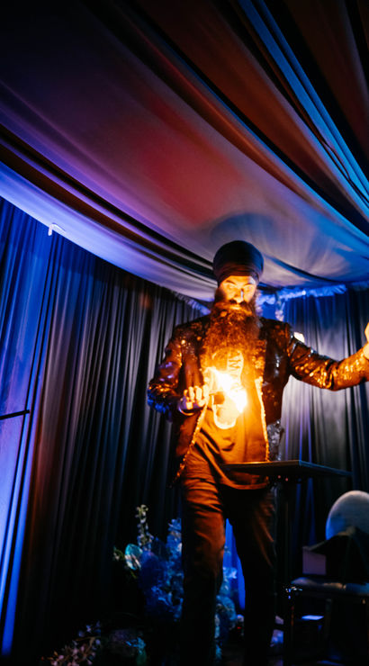 Stage performer with turban and long beard doing a fire trick while wearing a sequined jacket under dramatic blue and purple draped ceiling and curtains at an indoor event