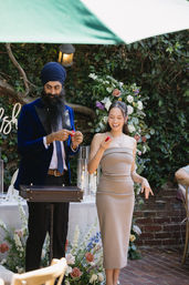 Garden wedding reception: man in a navy turban and velvet blazer performs a small magic trick with a red sponge ball while a smiling woman in a strapless beige dress watches by a floral arch and candlelit table.