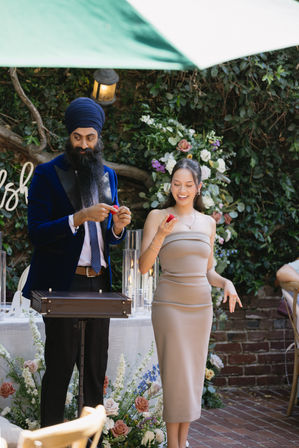 Garden wedding reception: man in a navy turban and velvet blazer performs a small magic trick with a red sponge ball while a smiling woman in a strapless beige dress watches by a floral arch and candlelit table.