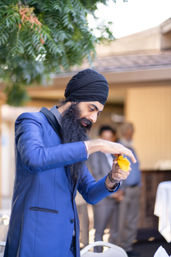 Man wearing a black turban and blue suit with a long beard carefully unfolding a yellow cloth or flower petal at an outdoor reception under a leafy tree.