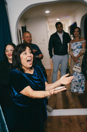 Woman in blue velvet gasps in delight while holding playing cards during a lively indoor house party, with guests watching in a hallway