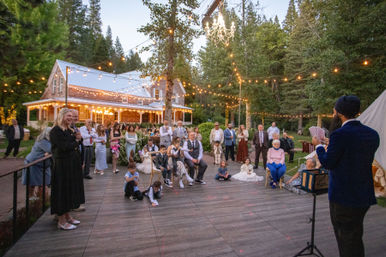 Evening outdoor wedding reception at a rustic farmhouse surrounded by trees, string lights and a hanging chandelier, guests gathered on a wooden dance floor watching a performer with playing cards.